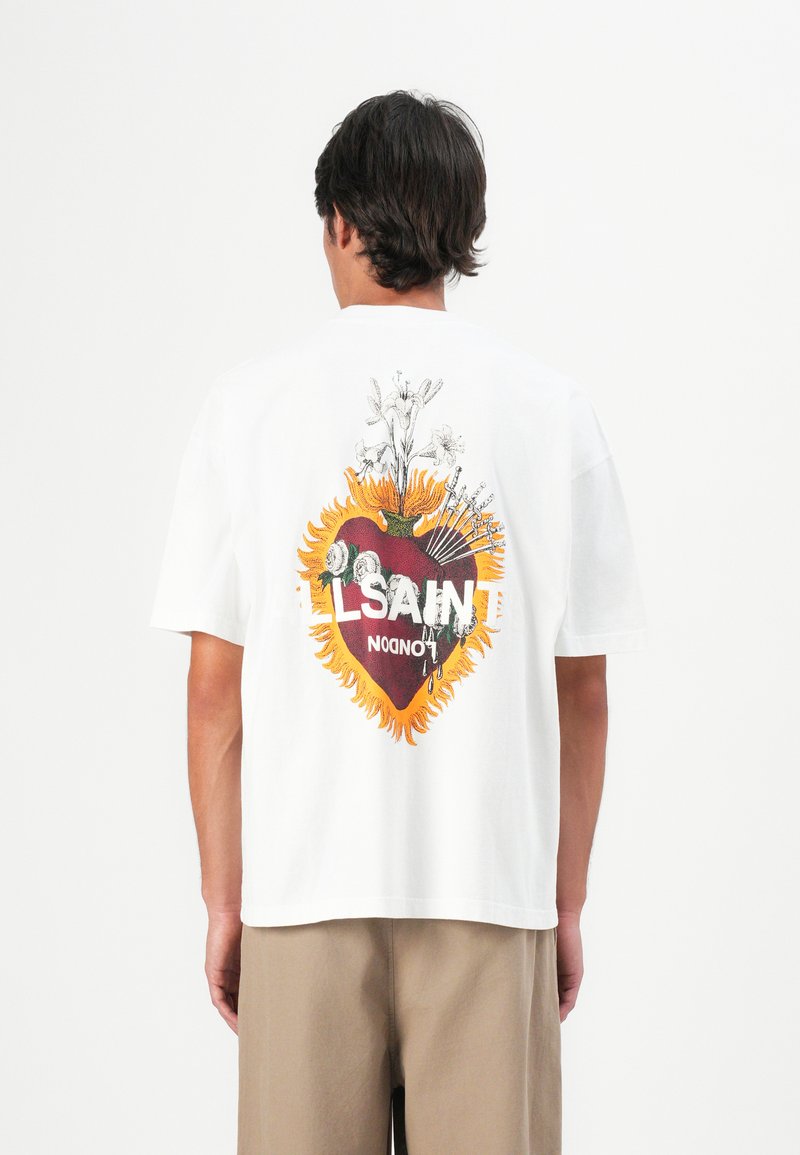 Man in een wit T-shirt met een grote afbeelding van een vlammenhart, witte bloemen en tekst "SAINT LONDON" op de rug, beige broek.