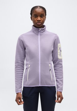 Młoda osoba z warkoczami, ubrana w jasnofioletową, miękką kurtkę na zamek i ciemne spodnie, stoi na tle jednolitego jasnego tła.