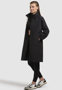 Cappotto nero alla lunghezza del ginocchio con collo alto, zip frontale e polsini elasticizzati. Texture liscia con tasche laterali, abbinato a leggings neri e sneakers.