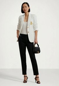 Mulher vestindo blaser branco, blusa branca, calças pretas, saltos altos pretos, segurando uma pequena bolsa preta, de pé contra um fundo simples.