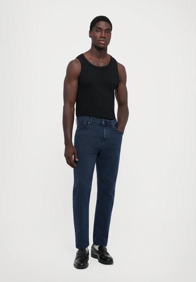 Man wearing black sleeveless top, dark blue jeans, and black dress shoes standing with one hand in pocket against plain background.