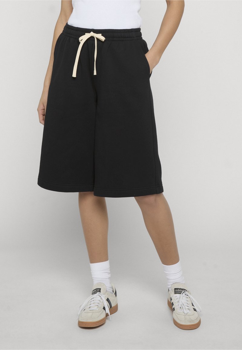 Personne portant un short noir mi-genou avec un cordon beige, des chaussettes blanches et des baskets Adidas blanches à rayures noires.