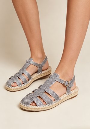 Pieds portant des sandales grises tissées à bout fermé avec des brides à la cheville et des semelles compensées en jute tressé sur un fond beige.