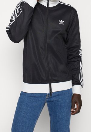 Personne portant une veste zippée noire Adidas avec des rayures blanches sur les manches et un jean bleu, main près du col, debout devant un fond blanc.