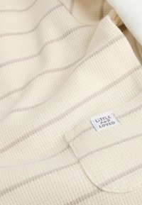 Tela color crema con sutiles rayas beige, que presenta un patrón de punto texturizado. Una pequeña etiqueta dice "PEQUEÑOS Y QUERIDOS."