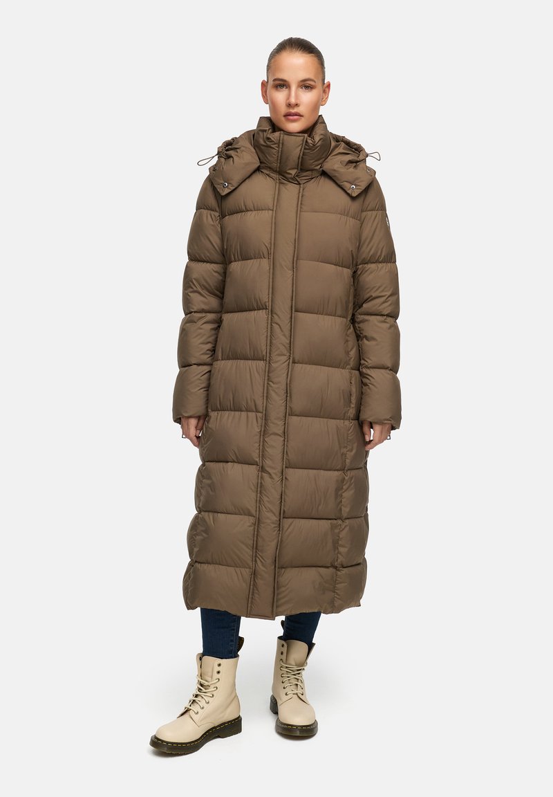 Langer, khakifarbener Steppmantel mit hohem Kragen, horizontalem Steppmuster und verstellbare Kapuze. Getragen über Jeans und cremefarbenen Schuhen im Kampfstil.