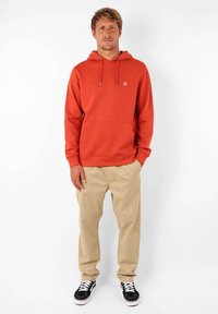 Sweatshirt à capuche rouge avec une poche avant, un cordon de serrage et un petit logo. Assorti avec un pantalon beige et des baskets noires avec des lacets blancs.