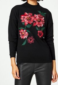 Svart stickad tröja med långa ärmar som har livfull broderi i röd och rosa blommor. Hög krage och strukturerad tyg.