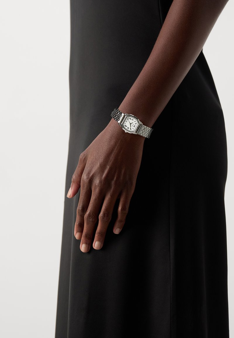 Montre en acier inoxydable avec un cadran octogonal, un cadran blanc, des index horaires noirs et un bracelet métallique texturé, portée sur une main contre une robe noire.