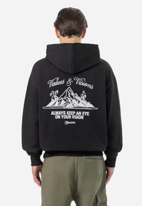 Person, die einen schwarzen Hoodie mit einem Berggrafik und dem Text "Values & Visions" sowie "Always keep an eye on your vision" auf dem Rücken trägt.