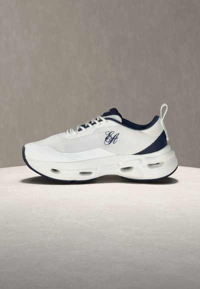 Baskets blanches et bleu marine avec fermeture à lacets à l'avant, semelle épaisse et rembourrée avec des poches d'air, et logo en script brodé sur le côté.