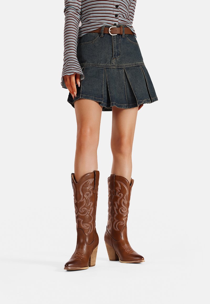 Stivali da cowboy in pelle marrone con cuciture decorative, punta appuntita e tacco a strati; indossati con una gonna di jeans e una maglietta a righe a maniche lunghe.