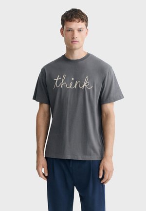 Jeune homme aux cheveux courts et bouclés portant un t-shirt gris foncé avec le mot "think" en cursive et un pantalon bleu marine, debout devant un fond uni.