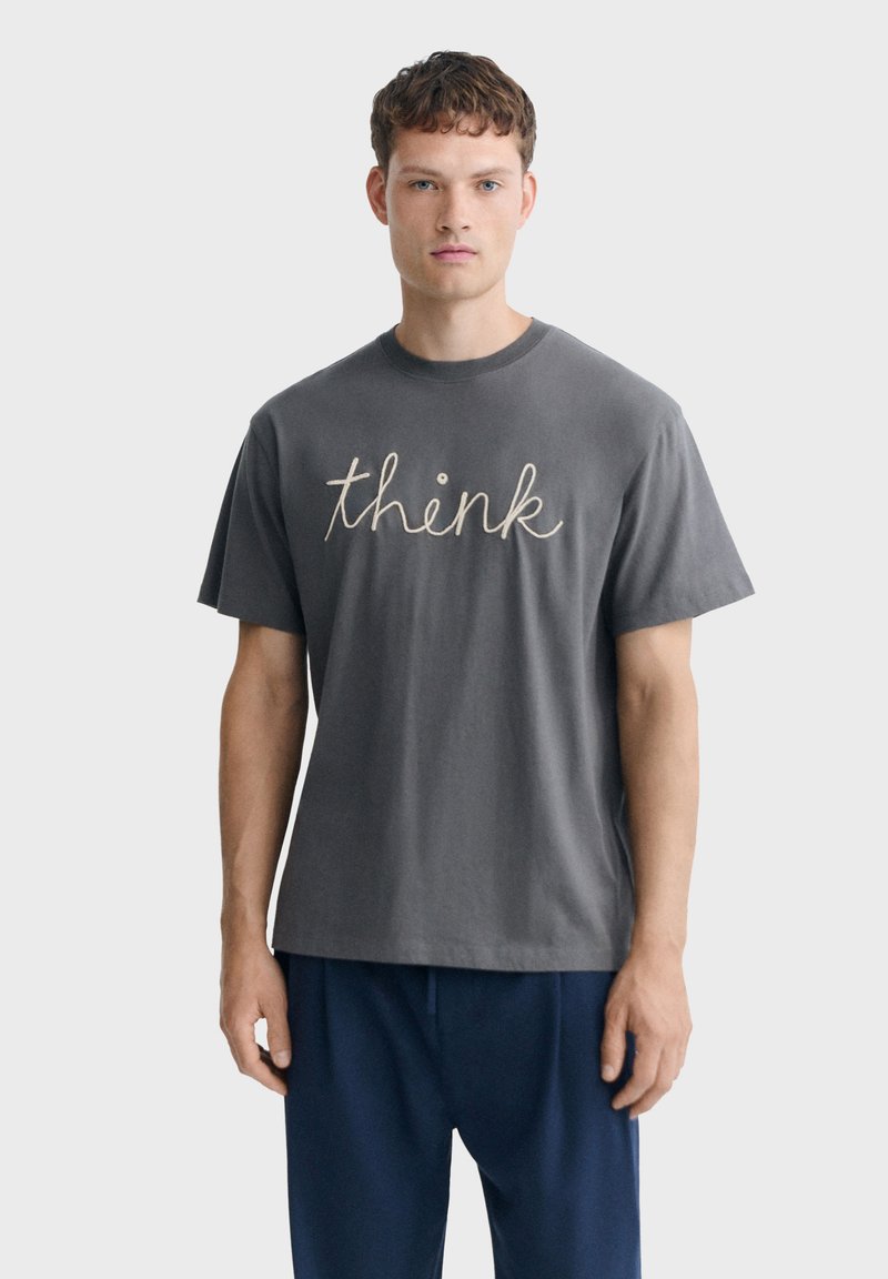 Jeune homme aux cheveux courts et bouclés portant un t-shirt gris foncé avec le mot "think" en cursive et un pantalon bleu marine, debout devant un fond uni.