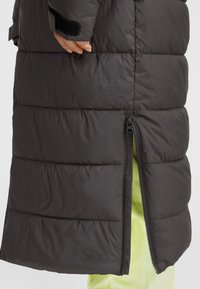 Svart pufferjacka med horisontella quiltade sektioner, en siddragkedja och en smidig textur. Insidan är klädd med ett klart gult tyg.