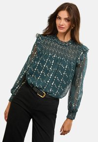 Türkiz áttetsző blúz arany pöttyökkel, fodros nyakkivágással, ráncolt testrésszel és hosszú, puffos ujjal. Fekete, magas derekú nadrággal párosítva.