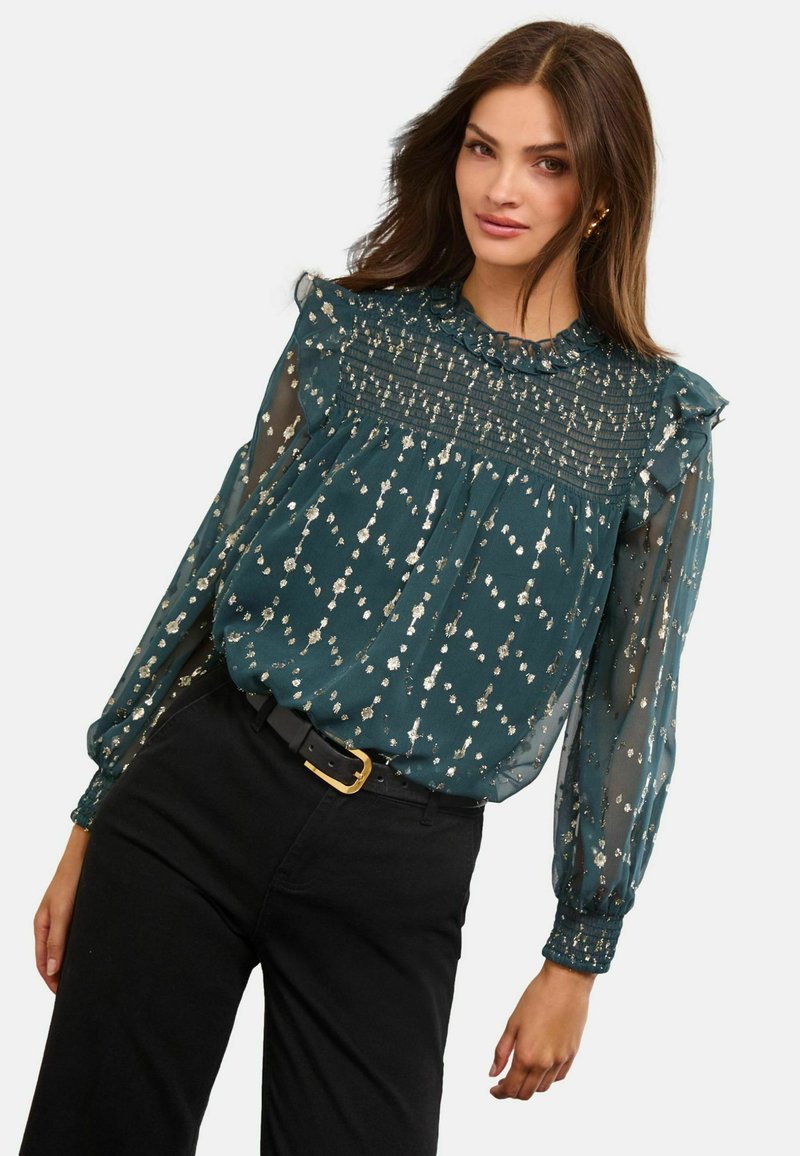 Türkiz áttetsző blúz arany pöttyökkel, fodros nyakkivágással, ráncolt testrésszel és hosszú, puffos ujjal. Fekete, magas derekú nadrággal párosítva.