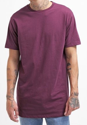 Homme portant un T-shirt violet uni à manches courtes et un jean bleu clair, montrant des tatouages sur le cou et les deux bras sur un fond blanc.