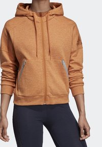 Sweat à capuche orange avec fermeture éclair, finition texturée, doté de deux poches avant zippées et d'une capuche. Comprend un logo discret sur la manche.