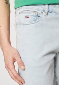 Vaqueros de denim azul claro con cinco bolsillos, un pequeño logo bordado en rojo, blanco y negro en el bolsillo trasero, y un diseño ajustado.