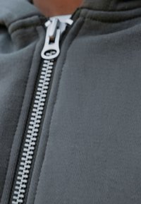 Tmavošedá mikina s hladkým, textúrovaným látkovým povrchom. Obsahuje výrazný strieborný zips, ktorý prebieha po prednej strane a zdôrazňuje jeho športový dizajn.