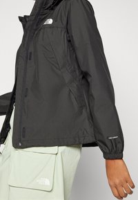 Musta vesitiivis takki, joka on valmistettu kevyestä materiaalista. Ominaisuuksiin kuuluvat elastiset hihansuut, vetoketjukiinnitys ja logot rinnassa ja hihassa.