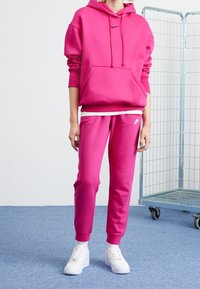 Felpa rosa con cordini e tasca frontale, abbinata a pantaloni rosa a sigaretta con logo. Sneakers bianche completano l'outfit.