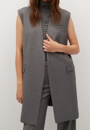 Kobieta ubrana w szarą kamizelkę bez rękawów na szarej bluzce z wysokim kołnierzem oraz dopasowane spodnie, stojąca na jednolitym tle.