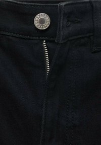 Gros plan sur la fermeture éclair d'un jean en denim noir partiellement ouverte sous un bouton argenté avec gravure de la marque.