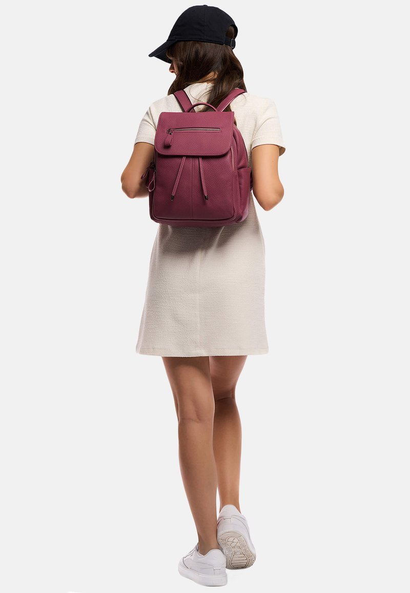 Sac à dos texturé violet avec des bretelles ajustables et une poche zippée à l'avant. Porté par une personne dans une robe légère et des baskets blanches.