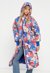 Cappotto imbottito floreale con cappuccio, caratterizzato da un mix di fiori rossi, blu e verdi su uno sfondo chiaro e polsini a coste blu in contrasto.