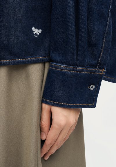 Χέρι δίπλα σε μπεζ παντελόνι που φοράει μανίκι σκουρόχρωμου μπλε τζιν μπουφάν με ορατή λευκή κεντητή πεταλούδα και καφέ ραφές.