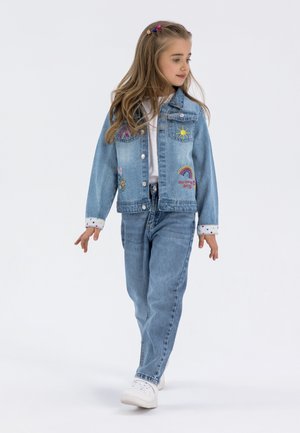 Jeans jakna z vezeninami mavrice in sonca, v kombinaciji s svetlo modrimi kavbojkami in belimi supergami. Ima manšete s pikami in klasičen kroj.