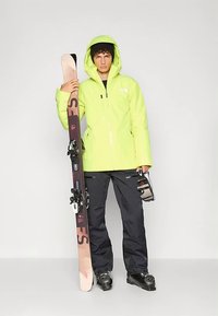 Hele-rohelise vettpidava jope, mustad suusapüksid, käes pruunid suusad ja mustad suusaprillid. Sile kangas koos kapuutsi ja tõmblukkudega pükstel.