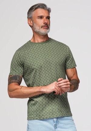 Mand med gråt hår og skæg iført grøn mønstret t-shirt og lyseblå jeans, som kigger til siden med hænderne foldet.