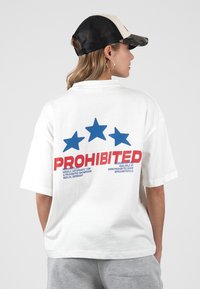 Άτομο που φοράει λευκό φαρδύ T-shirt με τρία μπλε αστέρια και κόκκινο κείμενο "ΑΠΑΓΟΡΕΥΕΤΑΙ" στην πλάτη, γκρι παντελόνι και μαύρο-άσπρο καπέλο.