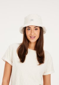 Witte bucket hat met een gestructureerd oppervlak, voorzien van een gestikt label aan de voorkant. Model draagt een bijpassende lichtgekleurde gestructureerde top.
