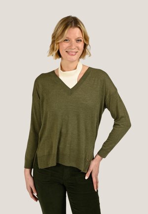 Femme souriante portant un pull col en V vert olive par-dessus un haut blanc, assorti à un pantalon vert foncé, debout avec une main sur la hanche.