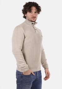Maglione lavorato a maglia beige chiaro con collo alto, dotato di una pattina con due bottoni scuri e polsini a coste. Indossato con jeans blu scuro.