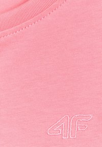 Gros plan sur un tissu rose avec col côtelé et logo brodé blanc "4F" sur le côté inférieur droit du textile.