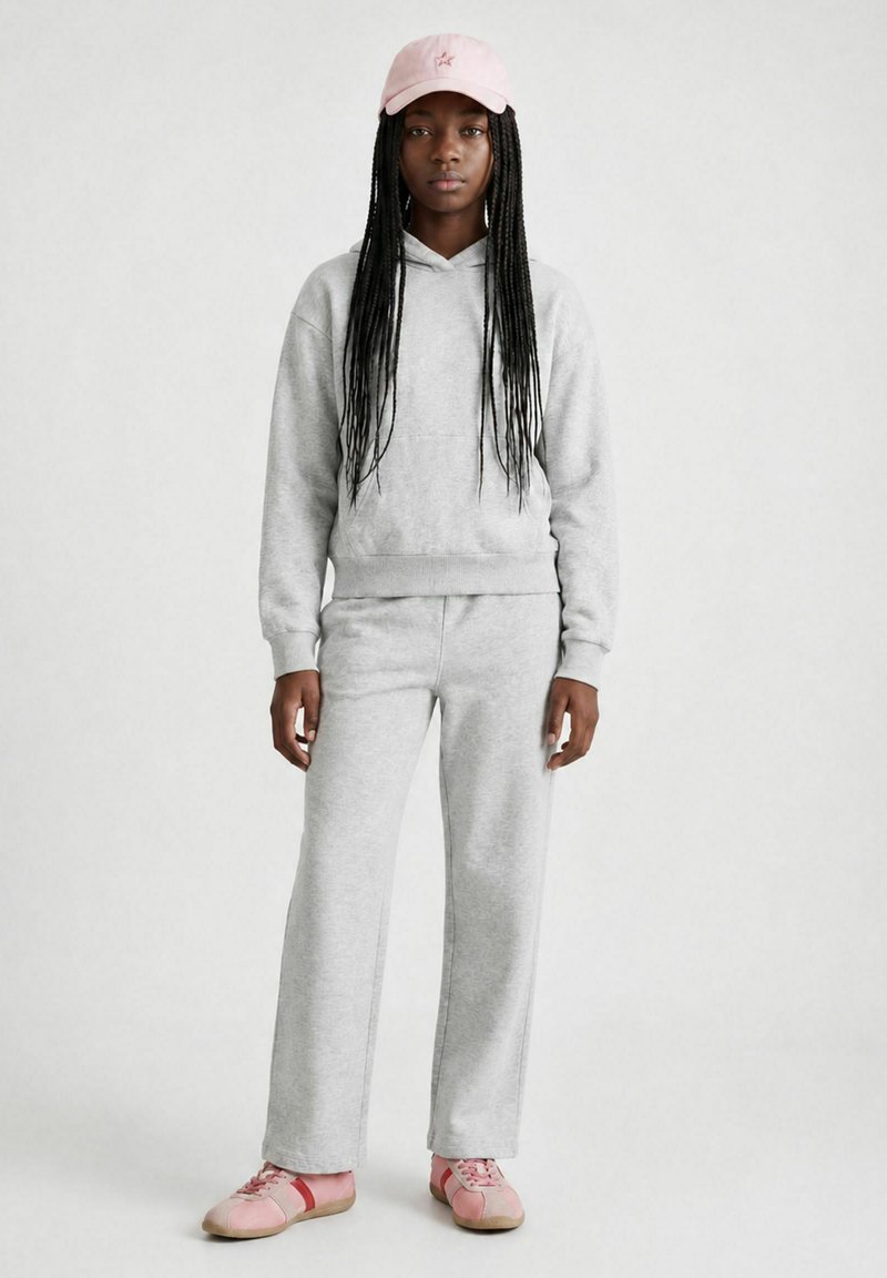 Jeune femme aux longues tresses porte un sweat à capuche gris clair et un pantalon, des baskets roses et une casquette rose clair, debout devant un fond uni.