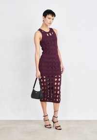 Robe en maille mauve avec des découpes circulaires, design sans manches, silhouette ajustée et longueur sous le genou. Le mannequin tient un petit sac noir.