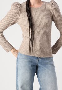 Beige ribbed knit sweater with puffed shoulders and long sleeves, paired with light blue high-waisted jeans. Textured, fitted design.