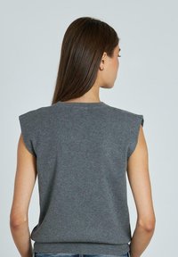 Suéter gris sin mangas con cuello redondo y hombros acolchados. Textura de punto suave con un ajuste ceñido en la cintura. Diseño minimalista.