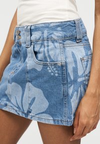 Τζιν μίνι φούστα με μεγάλο ανοιχτόχρωμο λουλουδένιο σχέδιο, μπροστινές τσέπες και διπλό κουμπί κλείσιμο, φορεμένη με λευκό τοπ.