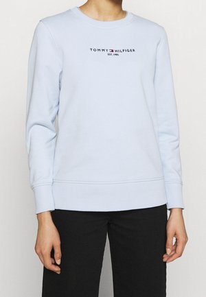 Osoba ubrana w jasnoniebieską bluzę Tommy Hilfiger z logo i napisem „EST. 1985”, w połączeniu z czarnymi spodniami, stojąca na jednolitym tle.