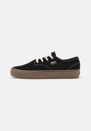 Zapatilla de ante negra con cordones blancos, suela de goma marrón, acento azul y un parche de logo en el lateral, con un diseño de tobillo bajo y puntera redondeada.