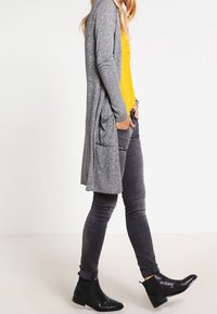 Lång grå cardigan med strukturerad stickning, öppen front och sidofickor. Matchas med en gul topp och svarta ankelskor.