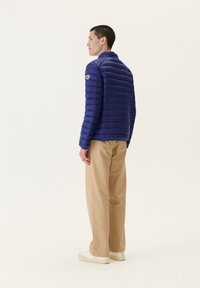 Chaqueta acolchada de marina hecha de material ligero con costuras horizontales, cuello alto y un parche de logo en la manga izquierda. Combinada con pantalones beige.
