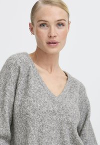Pelēks, pūkains, adīts džemperis ar V-veida kakla dizainu, ar platām piedurknēm un mīkstu tekstūru. Materiāls izskatās silts un ērts.